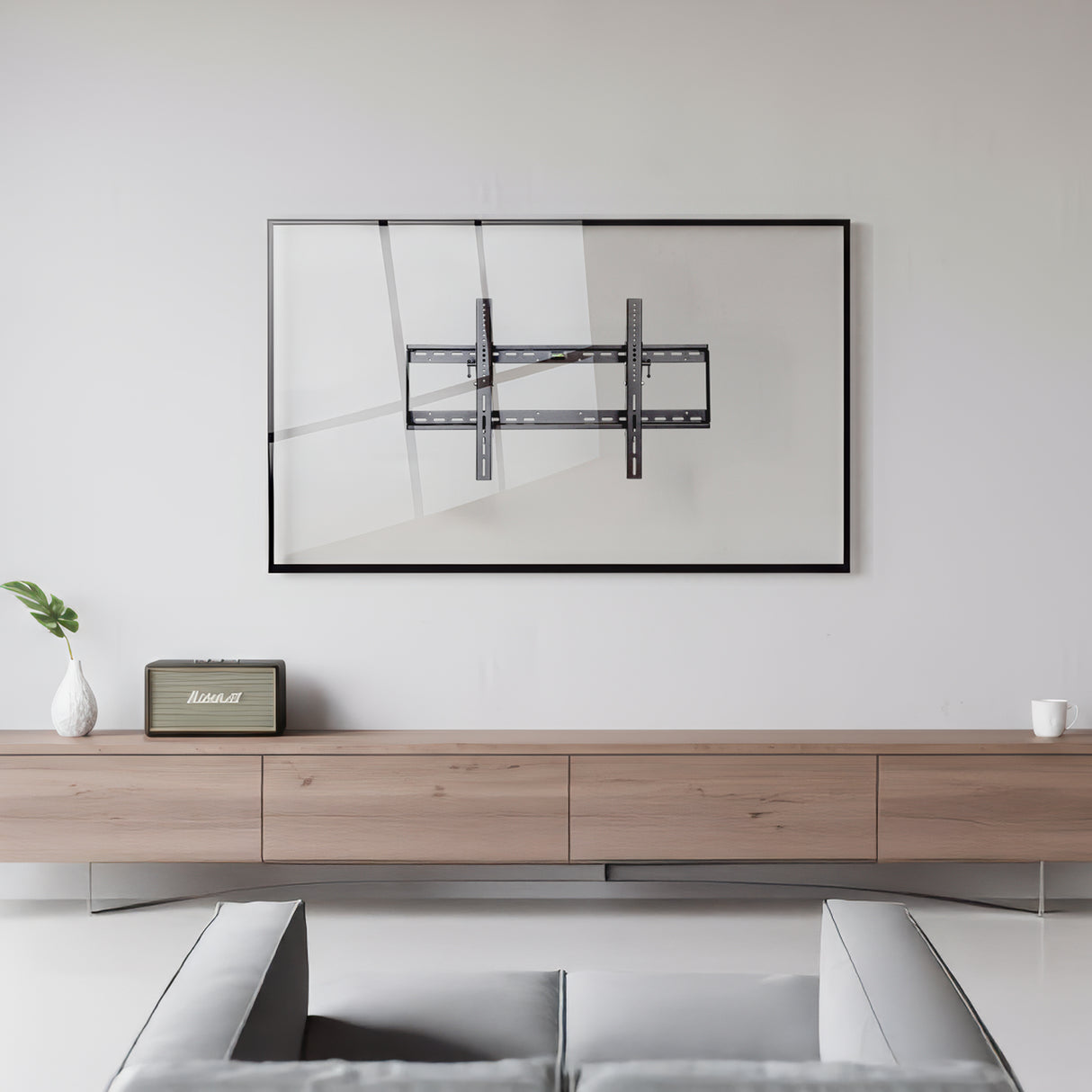 A minimalist living room features a Mount-It! Tilting TV Wall Mount with a slim, art-like frame on a white wall, above a light wood console. A small plant, retro radio, and cup decorate the console; part of a gray couch appears in front.