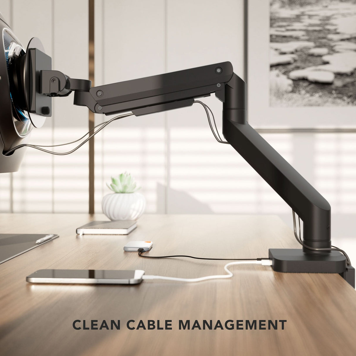 Heavy-Duty Monitor Mount with Gas Spring Arm The Mount-It! Heavy-Duty Monitor Mount with Gas Spring Arm holds a screen above a wooden desk, featuring clean cable management. A smartphone and plant are in the background. Text at bottom: CLEAN CABLE MANAGEMENT.