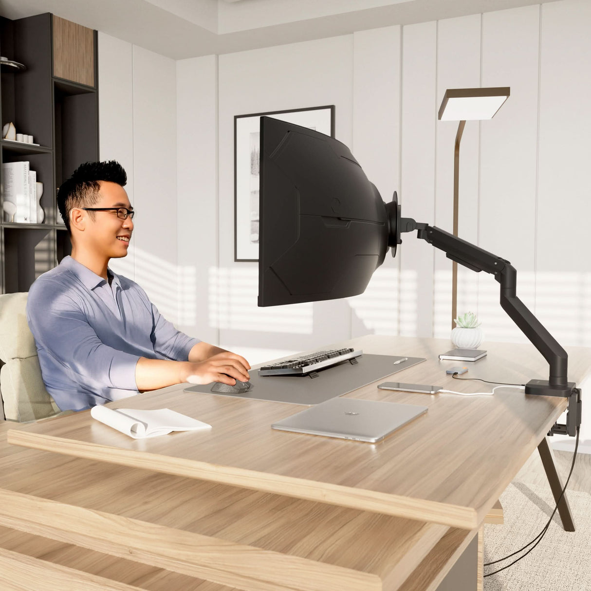 Heavy-Duty Monitor Mount with Gas Spring Arm A man sits at a modern desk in a bright office, smiling at a large monitor equipped with a privacy hood and the Mount-It! Heavy-Duty Monitor Mount with Gas Spring Arm. The tidy desk holds a keyboard, mouse, notepad, laptop, and small plant.