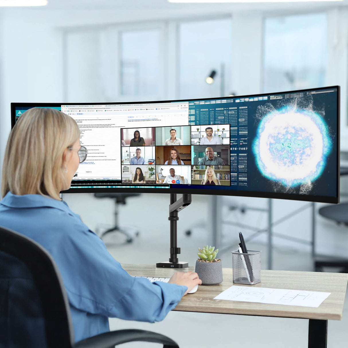 A person with short blonde hair works at a desk in an office, using a wide curved monitor held by the Mount-It! Heavy-Duty Monitor Mount with Pole & Gas Spring Arm. A keyboard, mouse, succulent plant, and pen holder are neatly arranged on the desk.