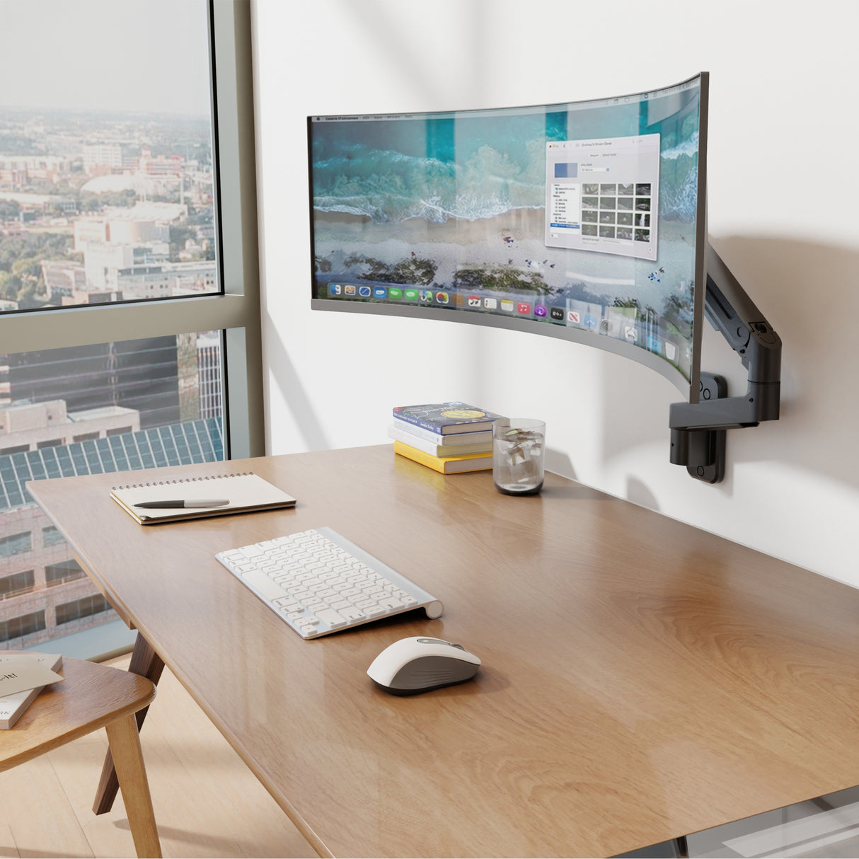 A modern office desk features an ergonomic workstation with a Mount-It! Heavy-Duty Single Monitor Wall Mount supporting a curved display, wireless keyboard and mouse, notebook, pen, and glass of water by a large window overlooking the cityscape.
