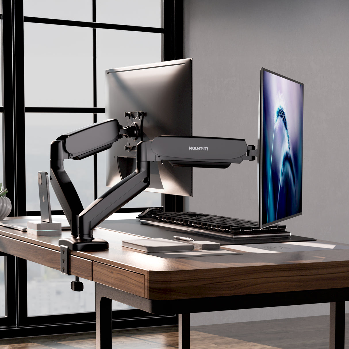 The Mount-It! Dual Monitor Desk Mount with Gas Spring Arms holds two screens side by side, offering full motion adjustability and smart cable management. The modern office setup includes a keyboard, mouse, notepad, and features large windows with a city view.