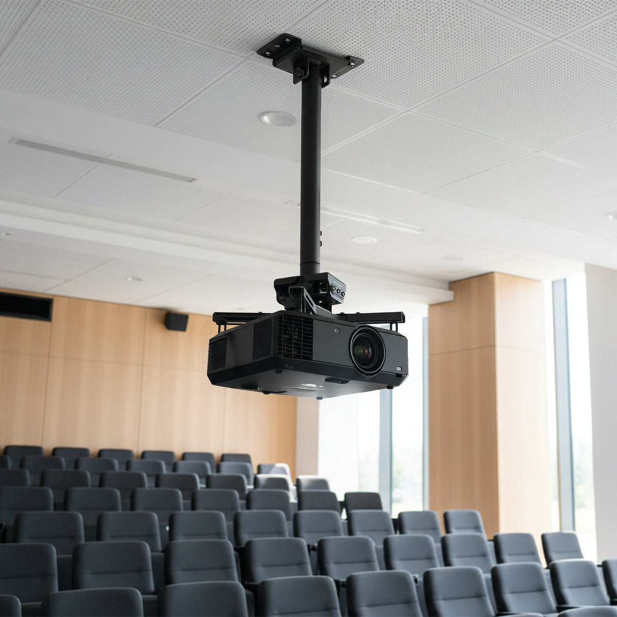 Heavy Duty Projector Ceiling Mount The Mount-It! Heavy Duty Projector Ceiling Mount holds a projector above rows of empty gray seats in a bright, modern lecture hall with light wood panels and large windows.