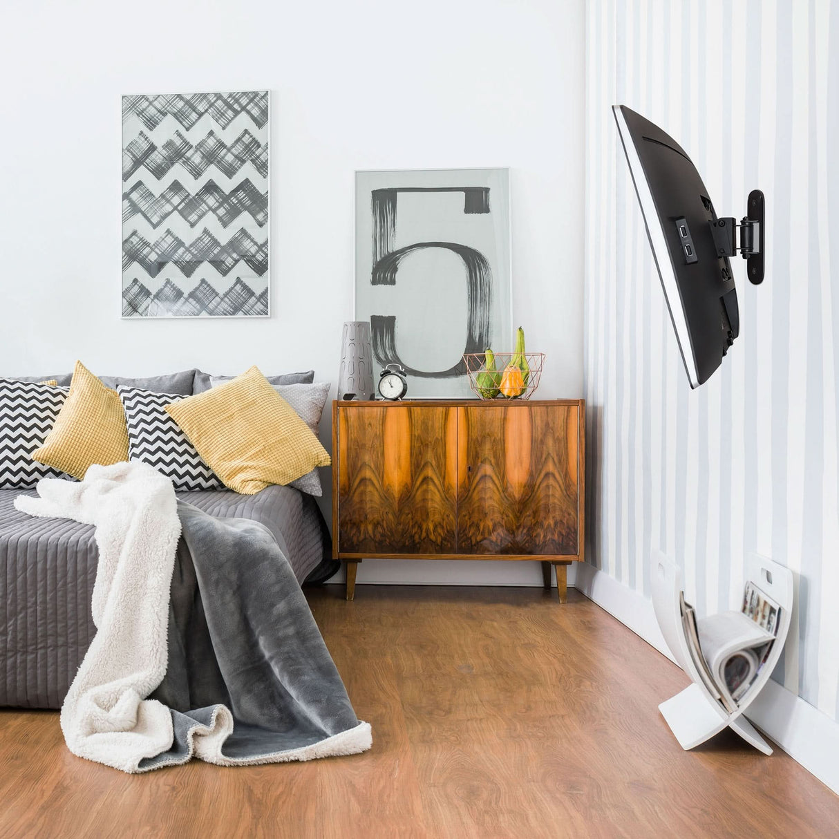 Single Stud Tilt and Swivel TV Wall Mount A modern living room with a gray sofa, yellow and gray cushions, a textured blanket, wooden sideboard with decor, abstract artwork, and a Mount-It! Single Stud Tilt and Swivel TV Wall Mount for the wall-mounted TV above the wood floor magazine holder.