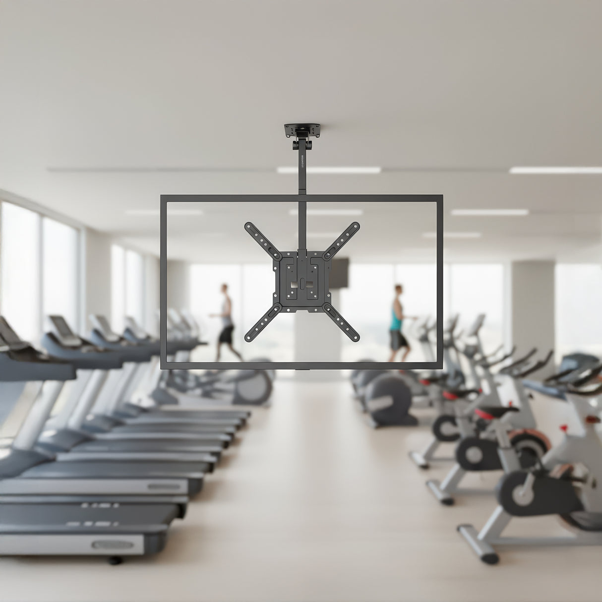 A Flip-Down Ceiling TV Mount by Mount-It! is installed in a modern gym with treadmills and exercise bikes. The mount is empty, with two people working out in the blurred background.
