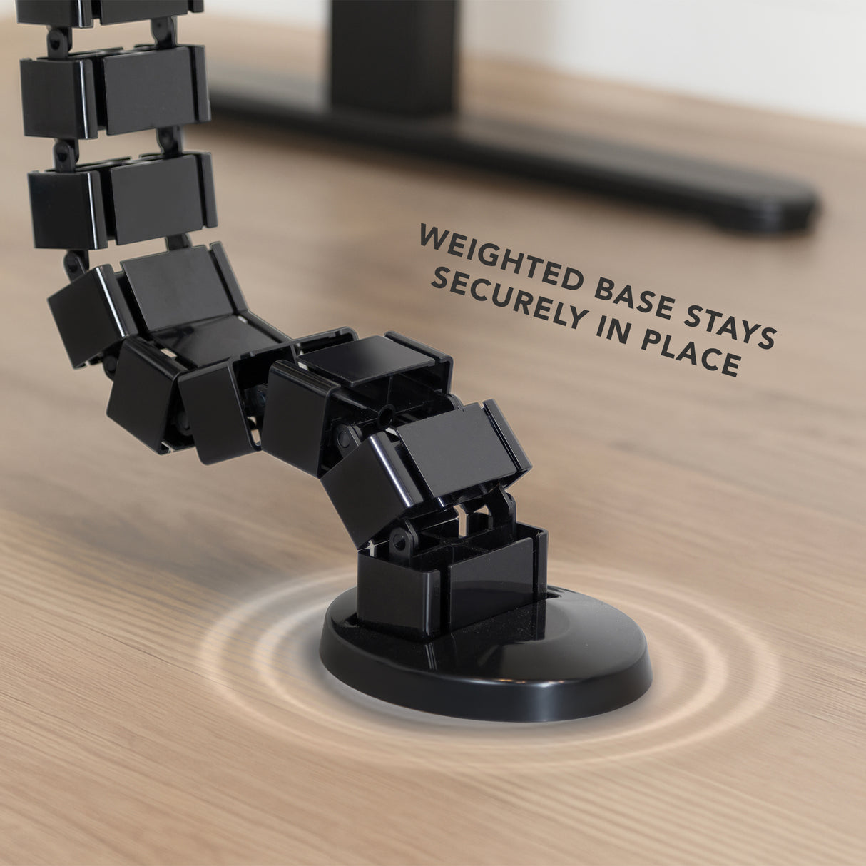 Cable Management Spine Close-up of the black Cable Management Spine featuring flexible snap-together segments with a weighted base resting securely on a wooden floor, designed to organize and protect cables while adjusting to desk height and providing strain relief.