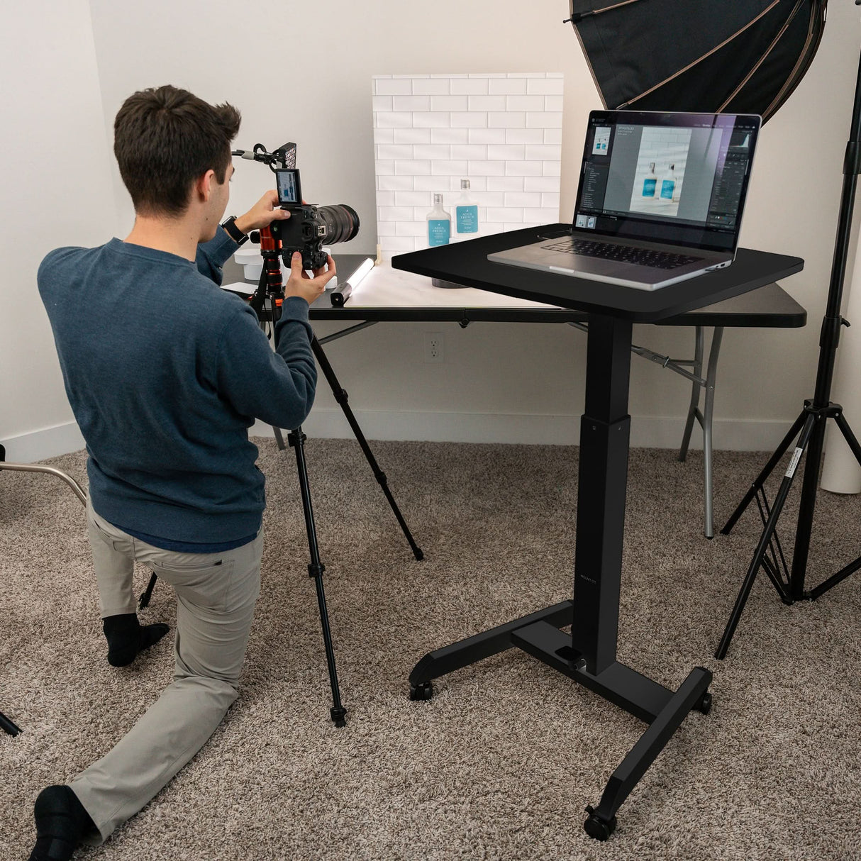 Height Adjustable Rolling Laptop Desk A man kneels on the carpet, photographing products on a table with a camera. Nearby stands the Mount-It! Height Adjustable Rolling Laptop Desk, and photography lighting equipment surrounds the workspace.