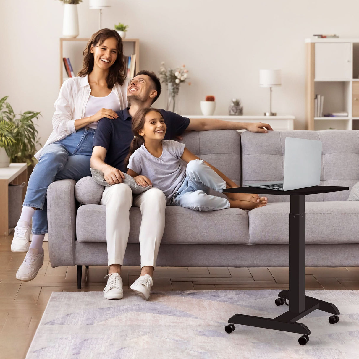 Height Adjustable Rolling Laptop Desk A smiling family of three sits on a gray sofa in a cozy living room, using the Mount-It! Height Adjustable Rolling Laptop Desk. The space features plants, a soft rug, and a light color scheme.