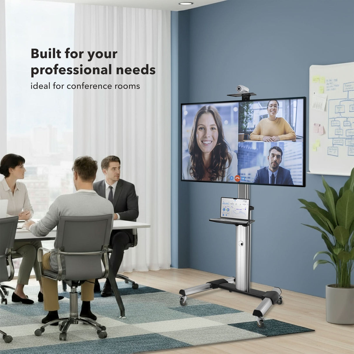 Heavy Duty Mobile TV Cart Three people sit at a table in a modern conference room as a video call displays on the Mount-It! Heavy Duty Mobile TV Cart with locking casters. Text: Built for your professional needs, ideal for conference rooms.
