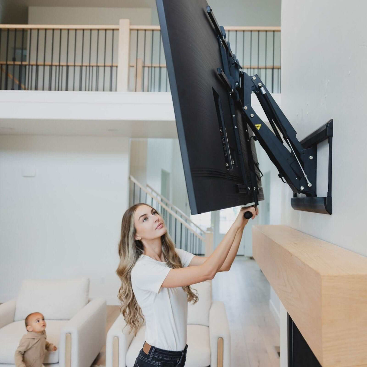 A woman installs the Mount-It! Pull Down Fireplace TV Mount above a fireplace, as a small child watches from the couch in a modern, bright living room.