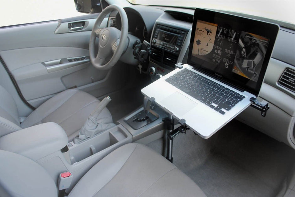 A laptop mounted on a stand inside a car, positioned over the passenger seat, with the dashboard and steering wheel visible. The laptop screen displays graphics and text.