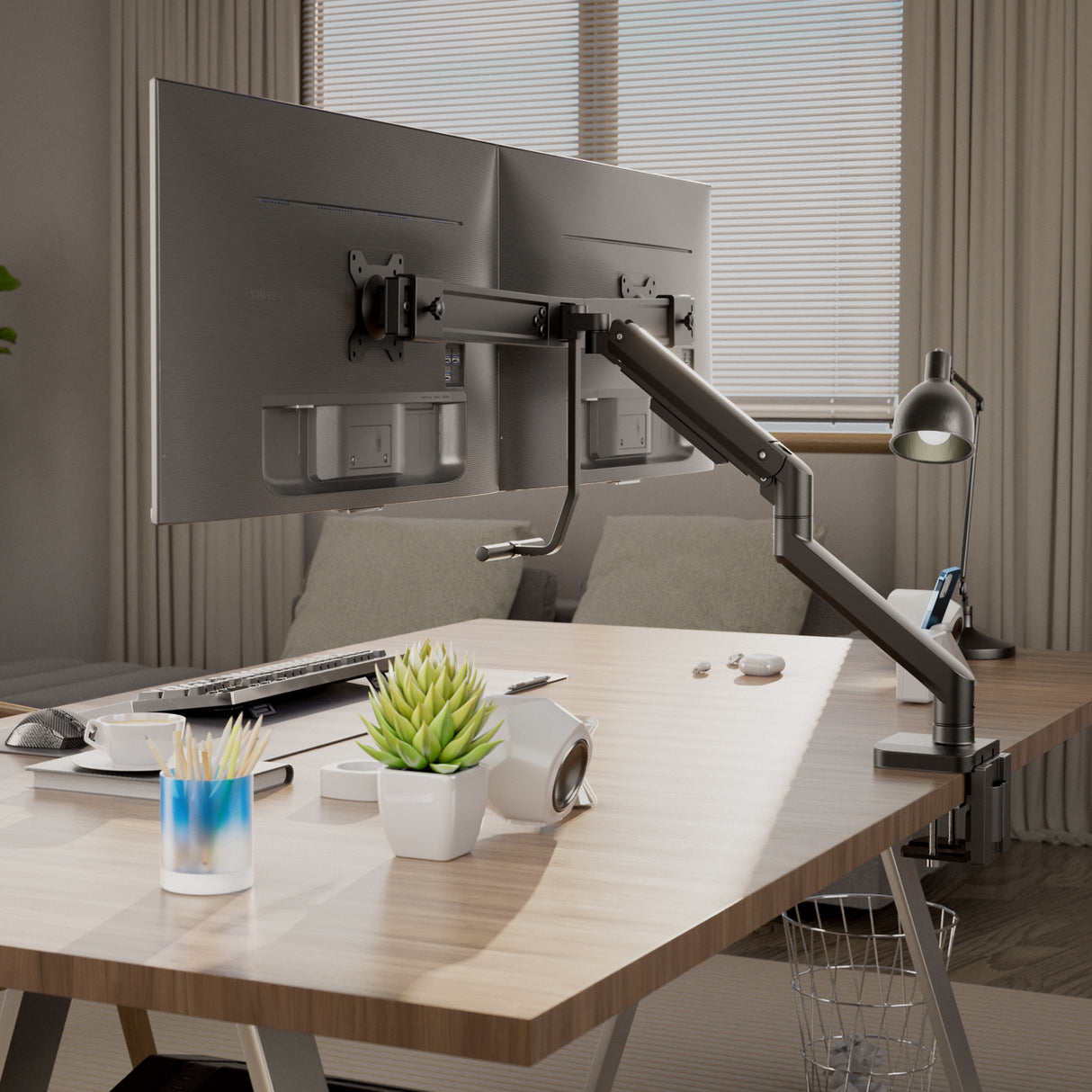 A modern workspace features a Mount-It! Dual Monitor Mount with Gas Spring Arm and Crossbar, plus a keyboard, mouse, lamp, potted plant, pen holder, and headphones neatly arranged on a wooden desk in front of window blinds.