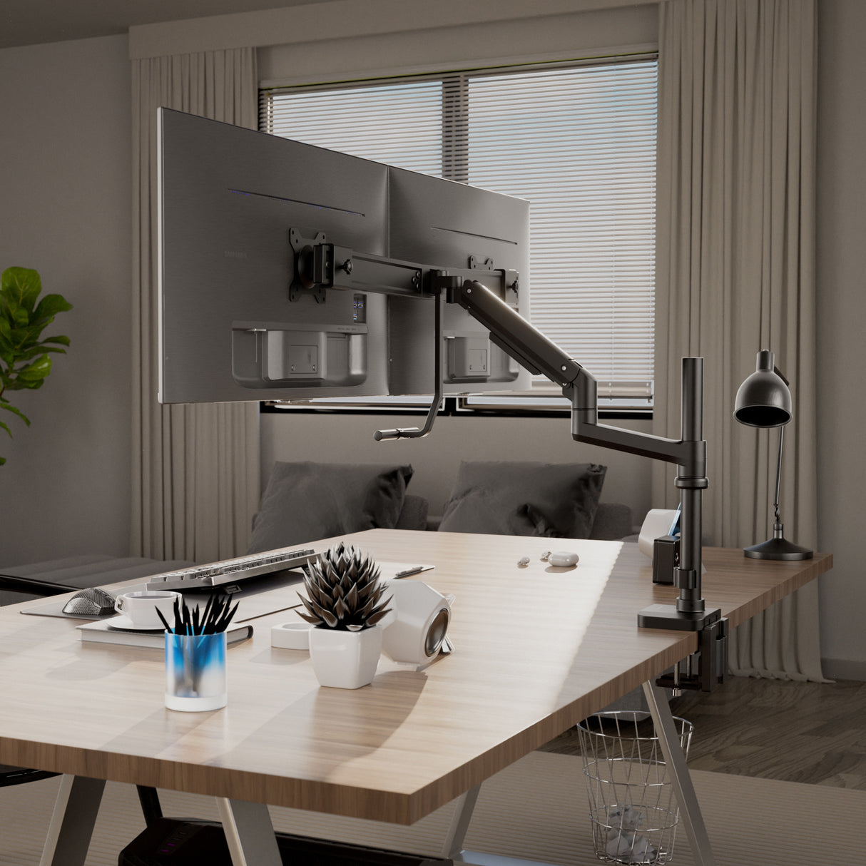A modern office desk features an ergonomic setup with the Mount-It! Dual Monitor Mount with Pole and Crossbar, adjustable USB-C monitor arm, keyboard, mouse, desk lamp, potted plant, VR headset, and supplies. A couch sits by a window with blinds.