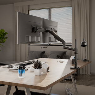 A modern office desk features an ergonomic setup with the Mount-It! Dual Monitor Mount with Pole and Crossbar, adjustable USB-C monitor arm, keyboard, mouse, desk lamp, potted plant, VR headset, and supplies. A couch sits by a window with blinds.