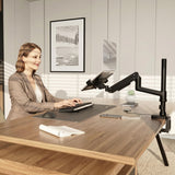 A woman in business attire types at a modern desk, using a tablet mounted on the Mount-It! Laptop Mount with Pole and Gas Spring Arm. Sunlight comes through window blinds, and a framed landscape photo hangs on the wall behind her.