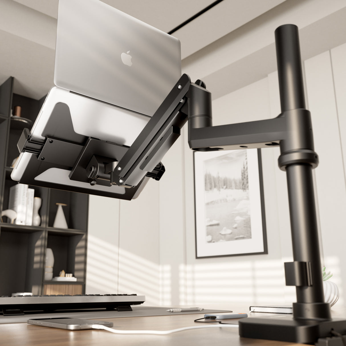 A close-up of a laptop on the Mount-It! Laptop Mount with Pole and Gas Spring Arm in a modern office, featuring an adjustable black desk arm, keyboard, mouse, and framed artwork in the background.