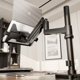 A close-up of a laptop on the Mount-It! Laptop Mount with Pole and Gas Spring Arm in a modern office, featuring an adjustable black desk arm, keyboard, mouse, and framed artwork in the background.
