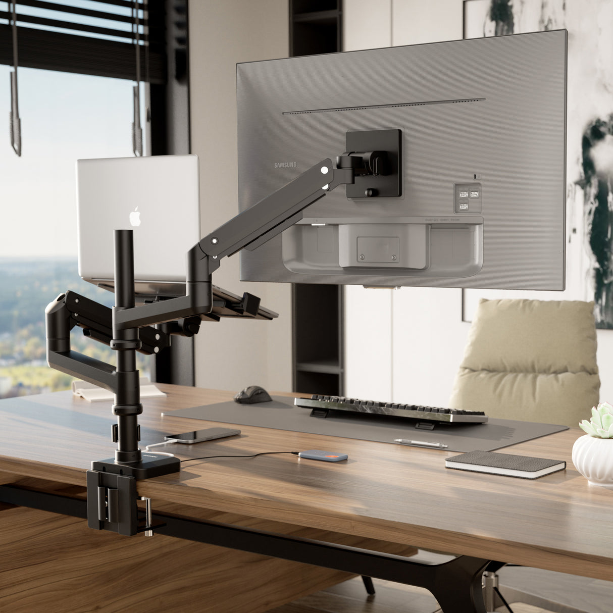 A modern office desk features the Mount-It! Heavy-Duty Monitor and Laptop Mount with Pole and Gas Spring Arms holding a large Samsung monitor and open MacBook, plus a keyboard, mouse, notebook, beige chair, and window view in the background.