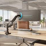 A Mount-It! Heavy-Duty Monitor Mount with Gas Spring Arm supports a large curved display on a modern wooden desk in a bright office with plants and large windows.