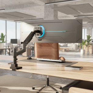 A Mount-It! Heavy-Duty Monitor Mount with Gas Spring Arm supports a large curved display on a modern wooden desk in a bright office with plants and large windows.