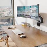 A modern office desk features an ergonomic workstation with a Mount-It! Heavy-Duty Single Monitor Wall Mount supporting a curved display, wireless keyboard and mouse, notebook, pen, and glass of water by a large window overlooking the cityscape.