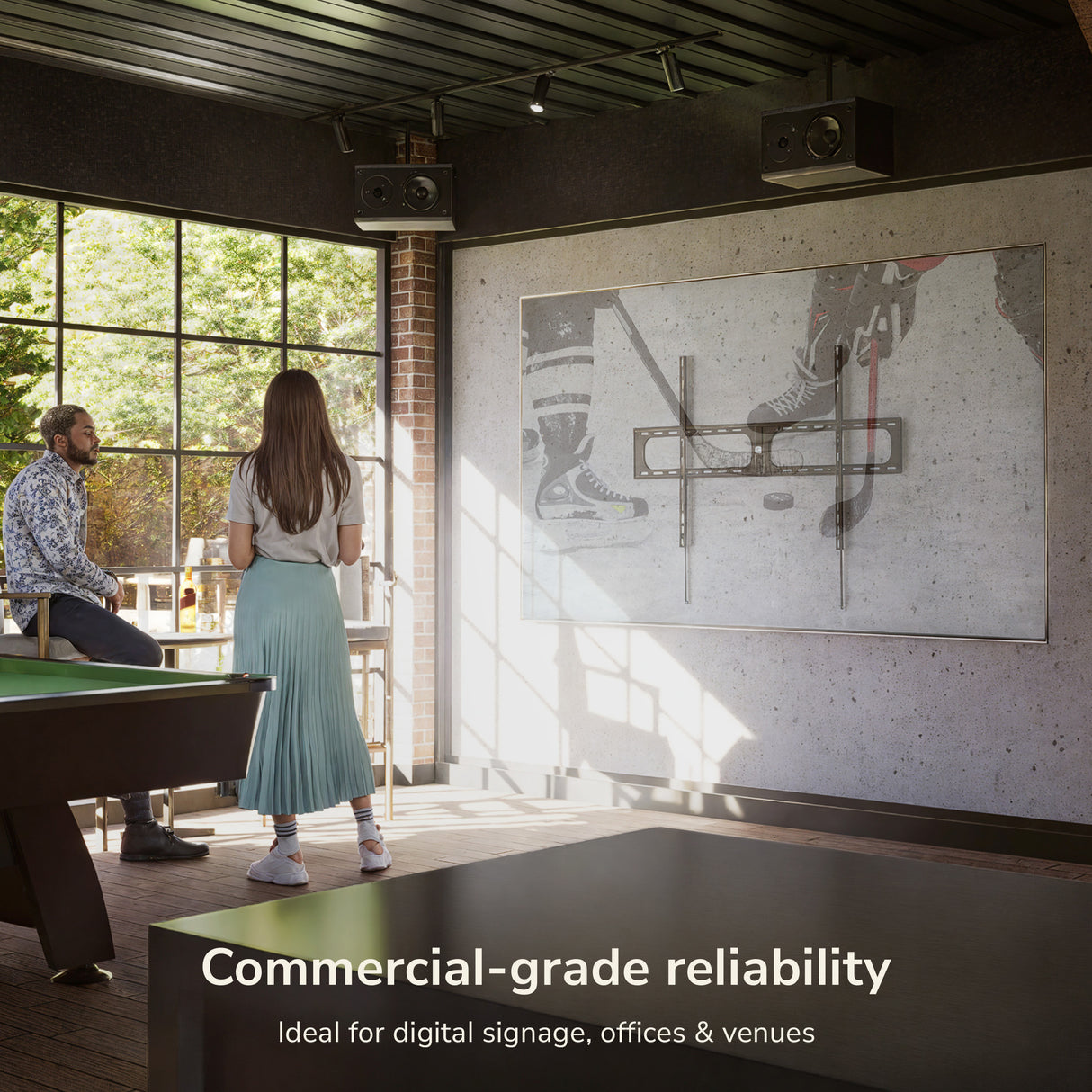 Two people stand in a modern room with large windows, watching hockey on a Mount-It! Heavy-Duty XXL Fixed TV Wall Mount. Text on screen: Commercial-grade reliability—ideal for digital signage, offices, and venues.