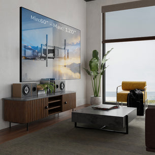 A large wall-mounted TV in a modern living room setting, shown above a wooden media console with speakers and decor. The room includes a gray coffee table, a yellow armchair by a large window, and a tall potted plant.