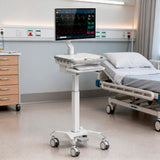 The Mount-It! MedHub Compact Mobile Workstation with locking casters and an adjustable workstation supports a monitor displaying vital signs beside a hospital bed in a bright, clean patient room.