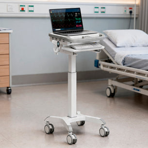A Mount-It! MedHub Scanner Holder on a mobile cart with a laptop showing patient vitals sits by a hospital bed. It includes a keyboard tray, scanner installation space, and four wheels for easy movement.
