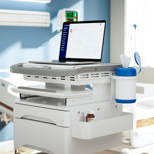 A Mount-It! MedHub Sanitizer Wipes Holder is shown with a laptop displaying medical records, a tissue dispenser, and medicine bottle on its tray; a hospital bed and medical monitor appear in the background along with storage for supplies.