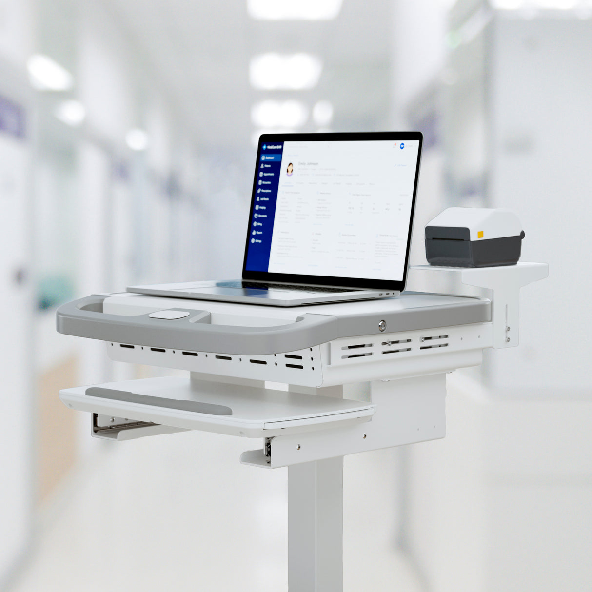 A Mount-It! MedHub Printer Holder mobile workstation with a laptop showing patient info and a compact printer on its shelf, set in a brightly lit hospital corridor.