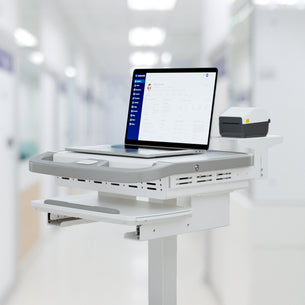 A Mount-It! MedHub Printer Holder mobile workstation with a laptop showing patient info and a compact printer on its shelf, set in a brightly lit hospital corridor.