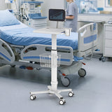 A hospital room with a bed and medical equipment features the Mount-It! MedHub Mobile Cart with Tablet Mount, cup holder, and wire basket on wheels beside the bed. In the background, a person in scrubs is walking.