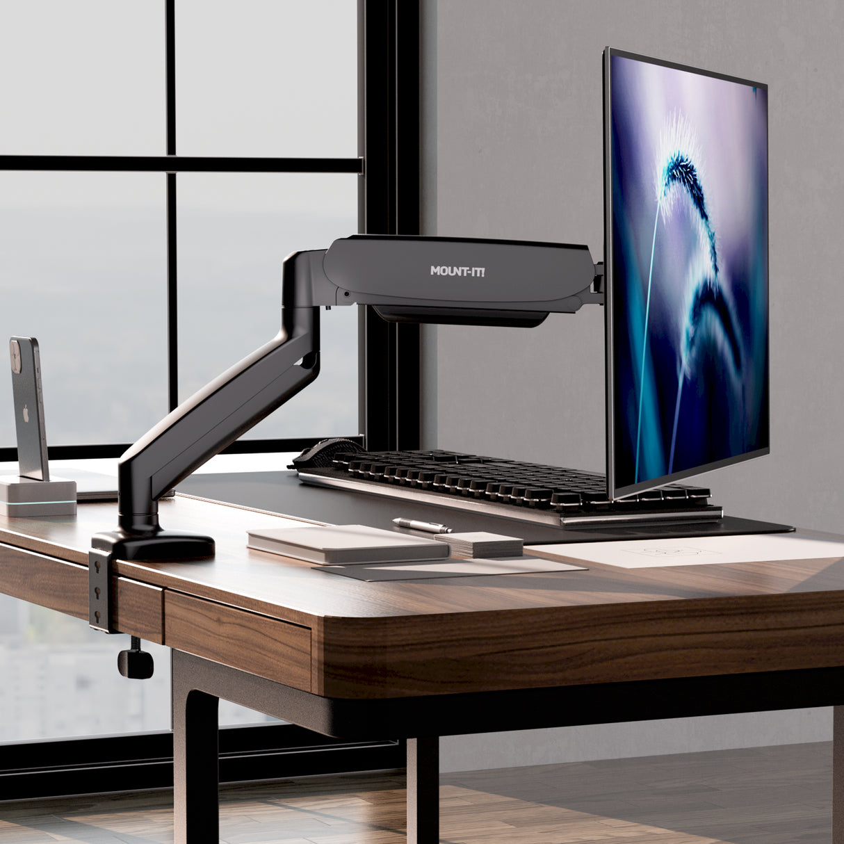 A modern office desk features the Mount-It! Single Monitor Desk Mount with Gas Spring Arm holding a slim monitor, plus keyboard, mouse, smartphone, notebook, pen, and desk organizer—all arranged by city-view windows with smart cable management.