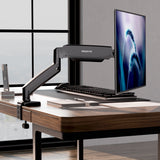 A modern office desk features the Mount-It! Single Monitor Desk Mount with Gas Spring Arm holding a slim monitor, plus keyboard, mouse, smartphone, notebook, pen, and desk organizer—all arranged by city-view windows with smart cable management.