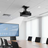A modern conference room features a Mount-It! Heavy-Duty Universal Projector Ceiling Mount, a large screen with charts, a wooden table, and black chairs. Tall windows with white vertical blinds fill the space with natural light.