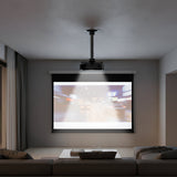 A modern living room features the Mount-It! Heavy-Duty Universal Ceiling Projector displaying a blurry city scene on a pull-down screen, facing two couches with a low table between them. Curtains cover the windows on one side.