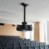 The Mount-It! Heavy Duty Projector Ceiling Mount holds a projector above rows of empty gray seats in a bright, modern lecture hall with light wood panels and large windows.