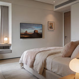 A modern, cozy bedroom features a neatly made bed with soft beige bedding, a Mount-It! Low-Profile Tilt TV Wall Mount displaying a desert scene, small framed art, a window seat with lamp, and soft natural light filtering in.