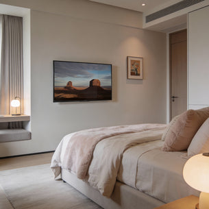 A modern, cozy bedroom features a neatly made bed with soft beige bedding, a Mount-It! Low-Profile Tilt TV Wall Mount displaying a desert scene, small framed art, a window seat with lamp, and soft natural light filtering in.