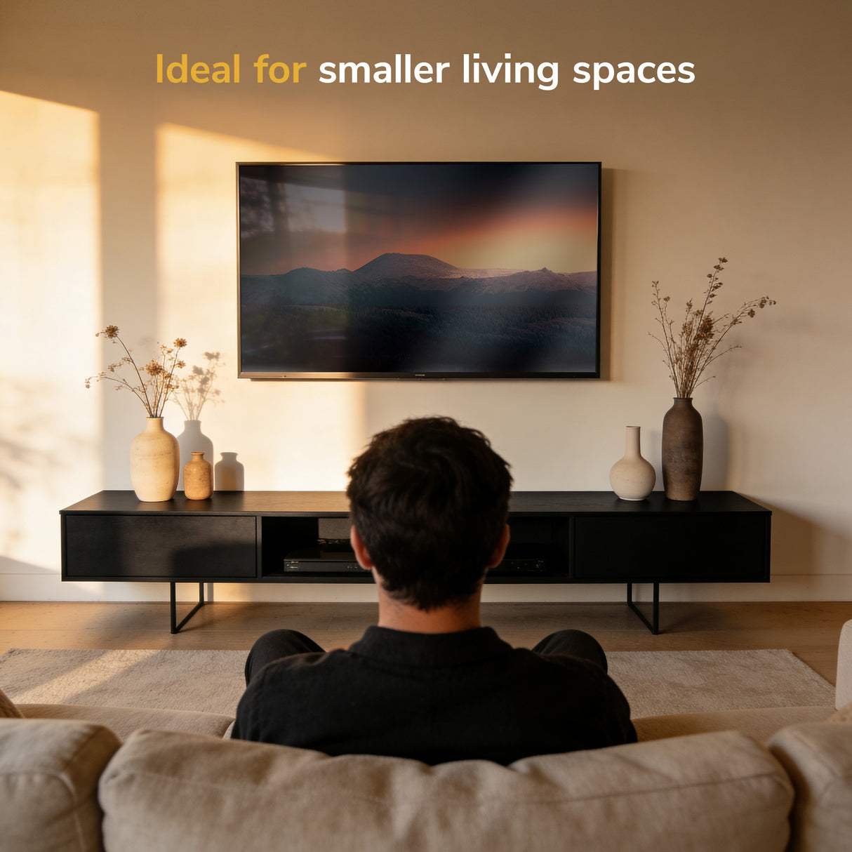 A person relaxes on a sofa facing a wall-mounted TV using the Mount-It! Low-Profile Tilt TV Wall Mount in a cozy living room with a black console, decorative vases, sunlight streaming in, and "Ideal for smaller living spaces" above the TV.