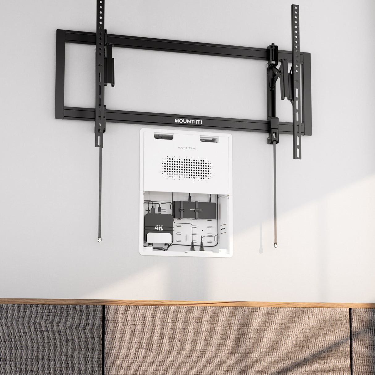 The Mount-It! In-Wall Media Box - Large is installed below a TV wall mount, organizing devices and cables neatly within the wall above a gray upholstered surface on a light-colored backdrop.
