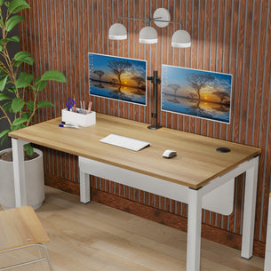 A modern workspace with a wooden desk features the Mount-It! Full Motion Dual Monitor Desk Mount (fits 32" monitors), dual screens displaying a sunset, white keyboard and mouse, desk lamp, pen holder, potted plant, and wood-paneled wall.