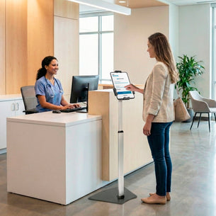 A woman uses a Mount-It! Secure Universal Tablet Floor Stand with Lock at a digital check-in kiosk in a modern medical office lobby, while the receptionist smiles from behind the front desk.