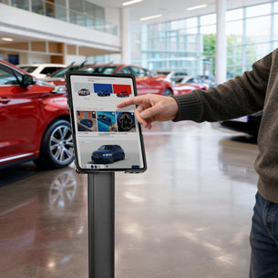 A person uses a Mount-It! Universal Tablet Floor Kiosk in a car showroom, browsing vehicle images and details on a secured touchscreen tablet, with several cars visible in the bright, modern space.