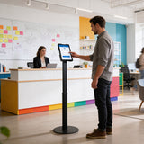 A man checks in using the Mount-It! Universal Tablet Floor Kiosk in a modern office lobby, while a woman works at a vibrant reception desk. The bright, open space is accented with notes and diagrams on the wall.