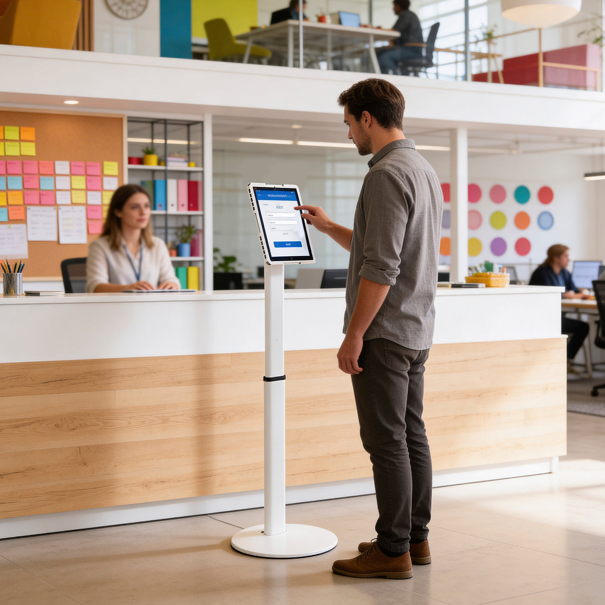A man uses the Mount-It! Universal Tablet Floor Kiosk at a digital check-in station in a modern office lobby, while a woman is at the reception desk. The background features colorful sticky notes and people working.