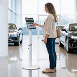 In a car showroom, a woman in a beige blazer, jeans, and flats uses the Mount-It! Universal Tablet Floor Kiosk as new cars are displayed in the background. She carries a purse over her shoulder while interacting with the secure tablet mount.