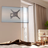 A Mount-It! Full Motion TV Wall Mount holds a flat-screen TV in a bright, modern living room with large windows, light curtains, a chair, side table with wine glass, and green plant. Sunlight streams through the cozy space.