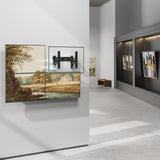 A contemporary art gallery features landscape paintings on the left, one split into four panels with a Mount-It! Pop-Out Video Wall Mount’s mechanical arm and pop-out design. Framed artworks are displayed on the right, with a bench in the background.
